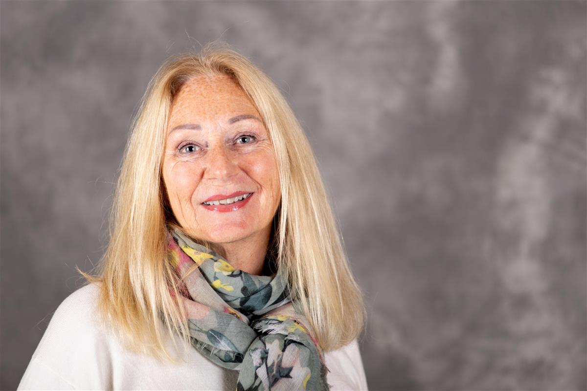 En kvinne med lyst hår og lyse klær smiler til kameraet foran en grå bakgrunn. - Klikk for stort bilde