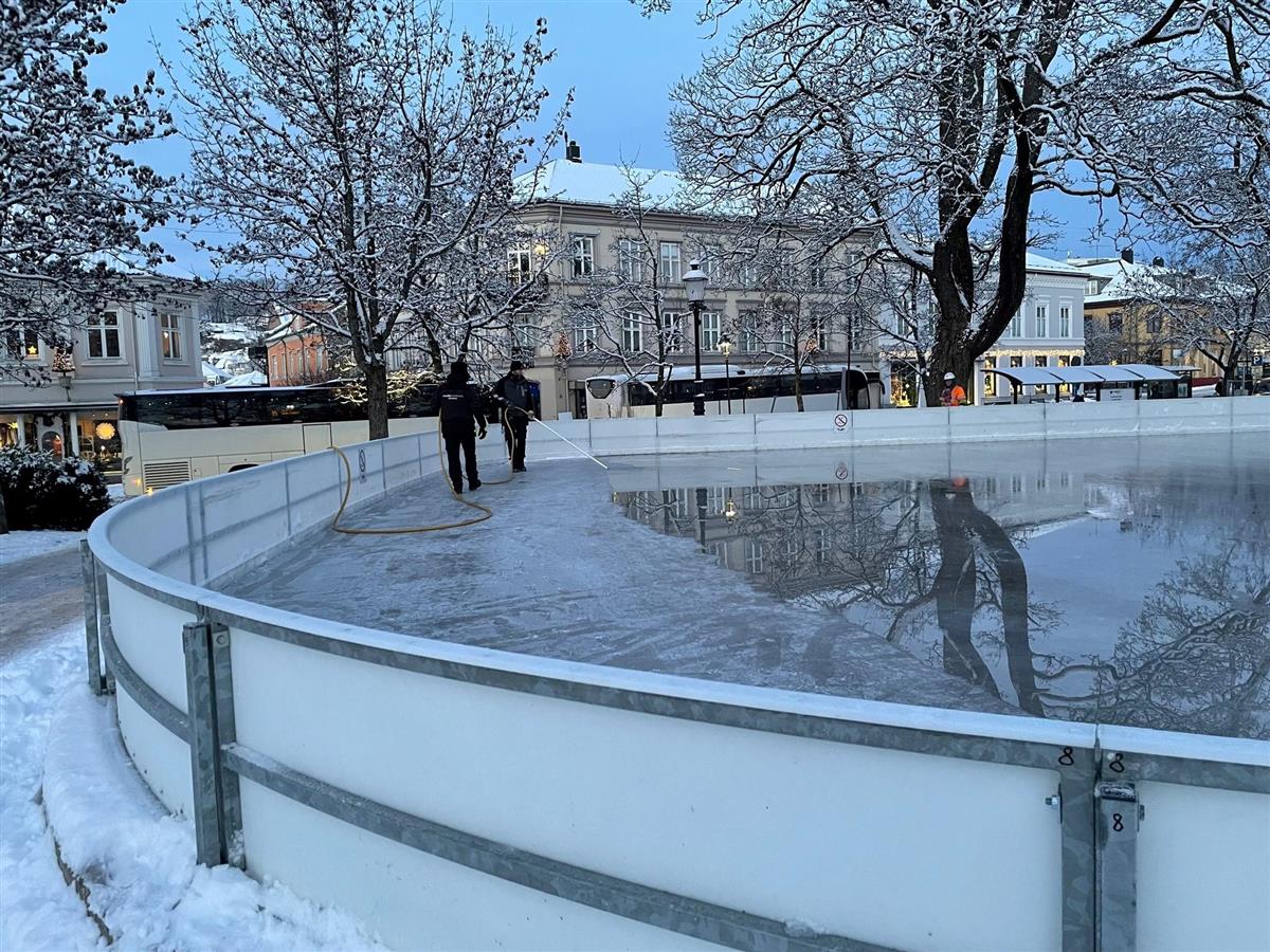 To personer fra Halden kommune jobber med islegging på skøytebanen i Busterudparken. Banen går rundt skulpturen Prillarguri som står på en høy granittsokkel midt på isen. - Klikk for stort bilde