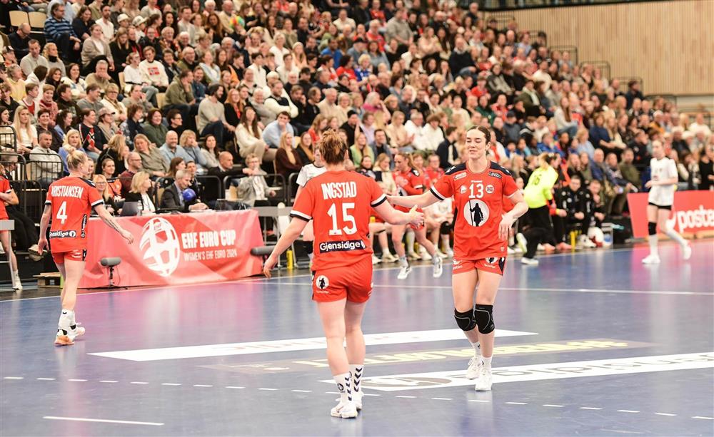 Håndballspillere på bane med mange tilskuere på tribune bak dem. - Klikk for stort bilde
