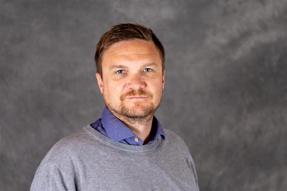 Mann med kort mørkt hår og grå blazer ser inn i kameraet - Klikk for stort bilde