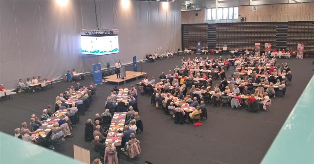 Mange mennesker sitter ved mange langbord i en hall - Klikk for stort bilde