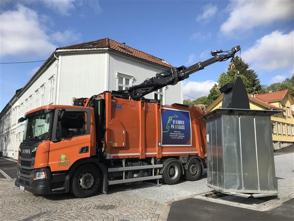 Oransje søppelbil løfter en nedgravd container i ei gate med gatesten