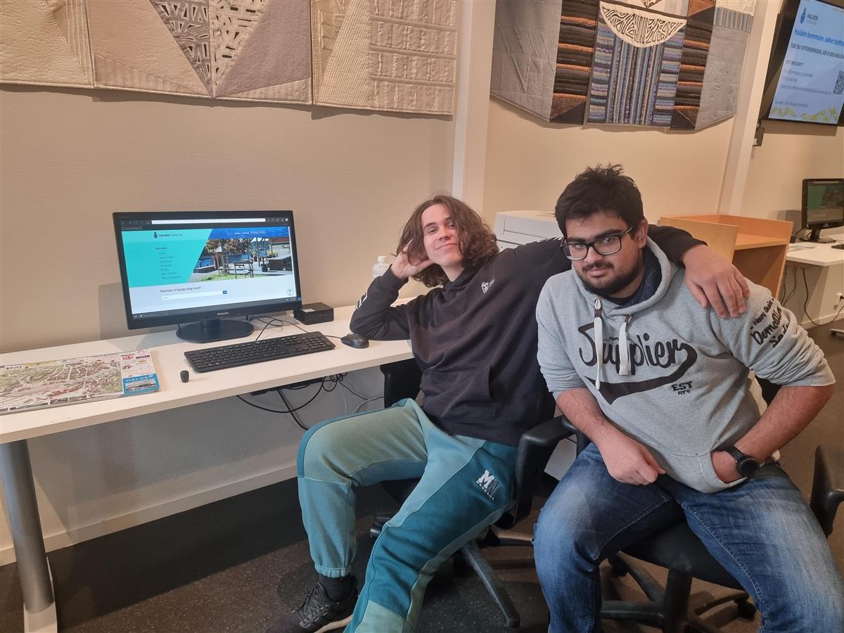 Elevene Håkon Gjøsund og Abdul Butt sitter ved siden av en stasjonær PC i serviceområdet til Halden kommune. Her skal de hjelpe folk med digitale vansker. - Klikk for stort bilde