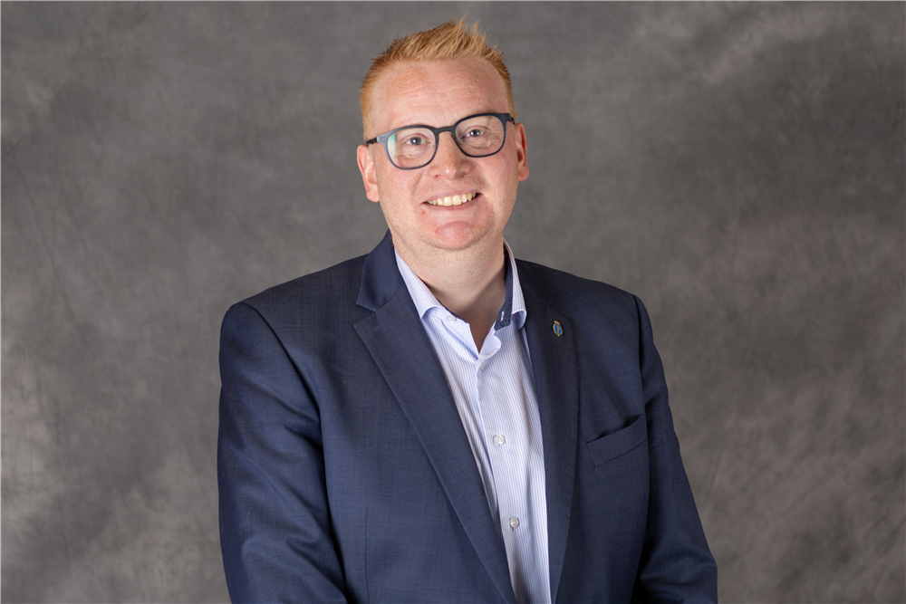 Mann med kort oransje hår og briller har på seg blå dressjakke foran en grå bakgrunn - Klikk for stort bilde