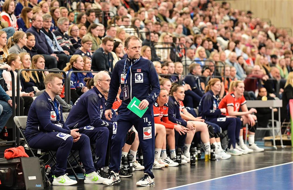 Håndballspillere og håndballtrenere på benk med mange tilskuere på tribune bak dem. - Klikk for stort bilde