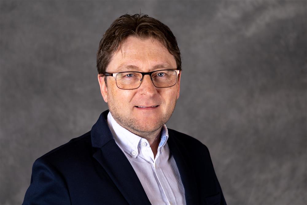 Mann med kort mørkt hår og briller, har på seg mørkeblå genser og hvit skjorte ser inn i kameraet - Klikk for stort bilde