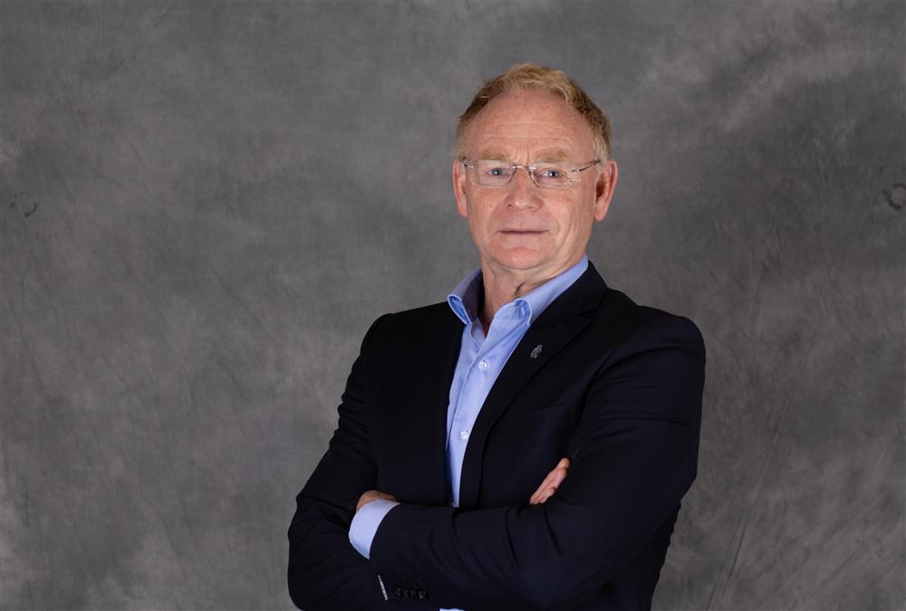 Mann med kort hår og lyse briller , har på seg en dressjakke med blå skjorte under, står foran en grå bakgrunn - Klikk for stort bilde