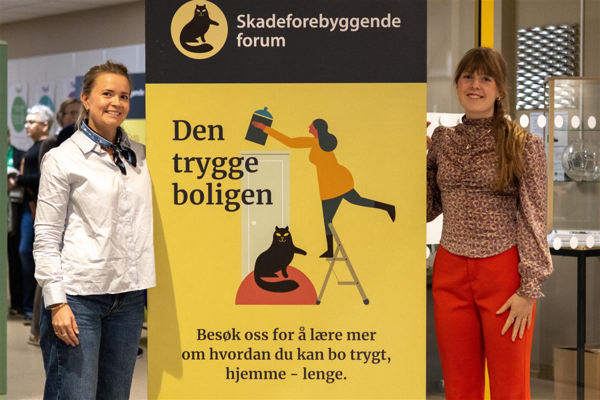 To kvinner står på hver sin side av en stor plakat som illustrerer utstillingen "Den Trygge Boligen". - Klikk for stort bilde