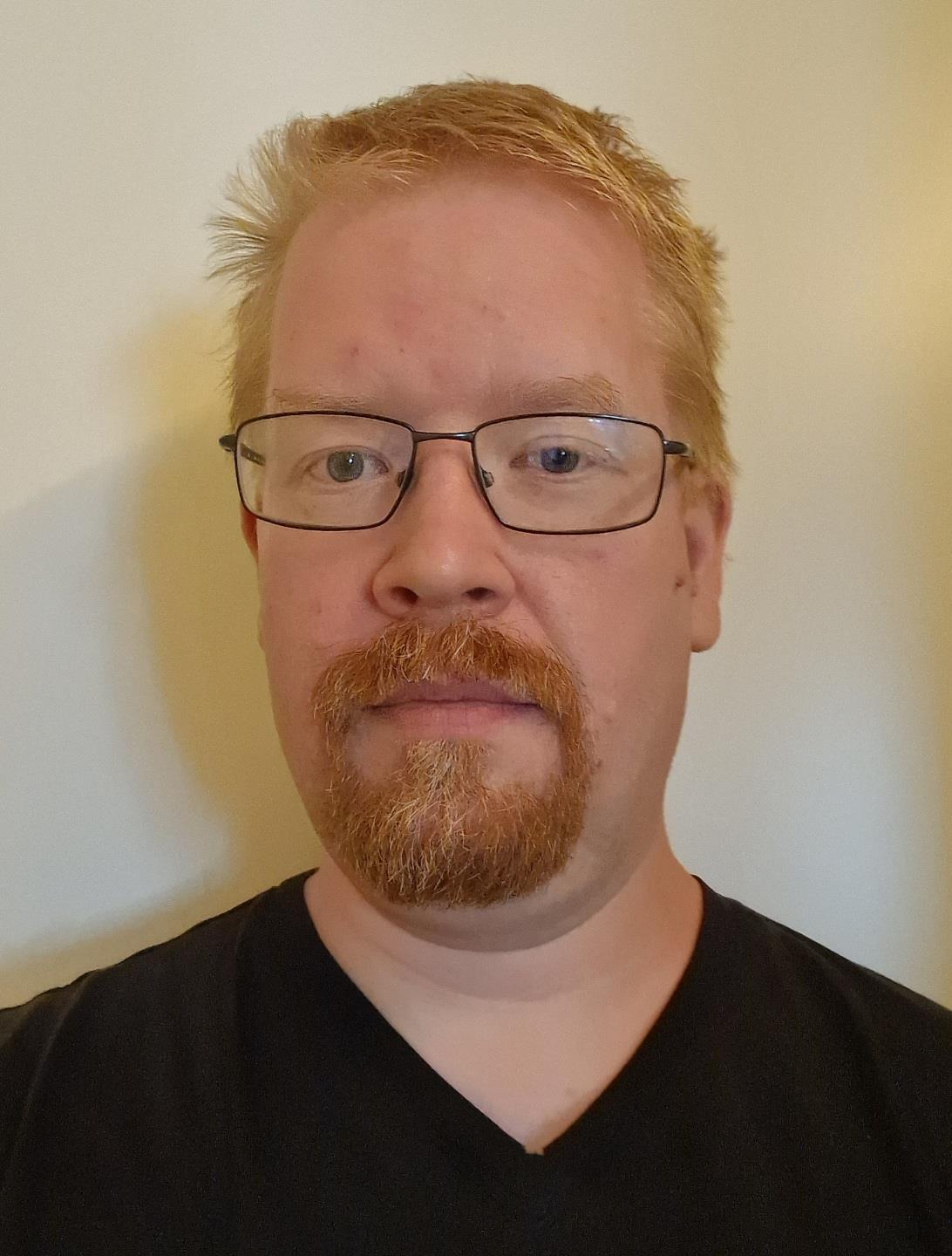 En mann med kort rødt hår og skjegg, sorte briller og svart t-skjorte. - Klikk for stort bilde