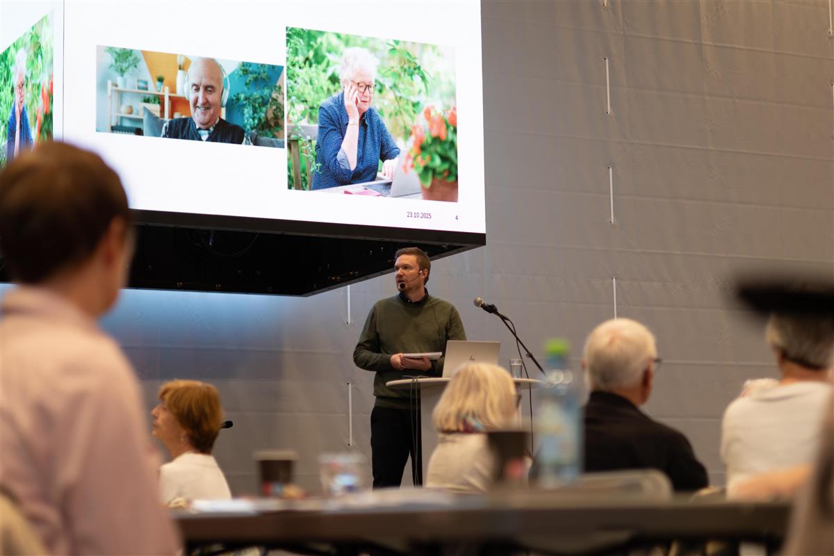 En mann står bak et podium og snakker til et stort publikum av eldre mennesker. - Klikk for stort bilde