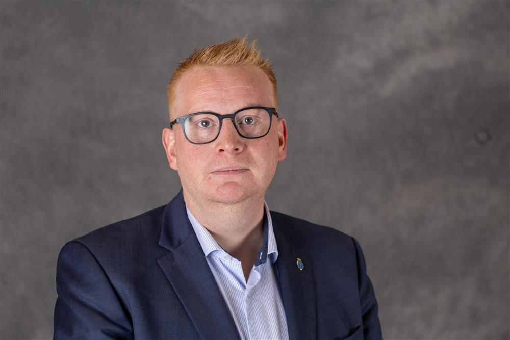 Mann med kort oransje hår og briller har på seg blå dressjakke foran en grå bakgrunn - Klikk for stort bilde