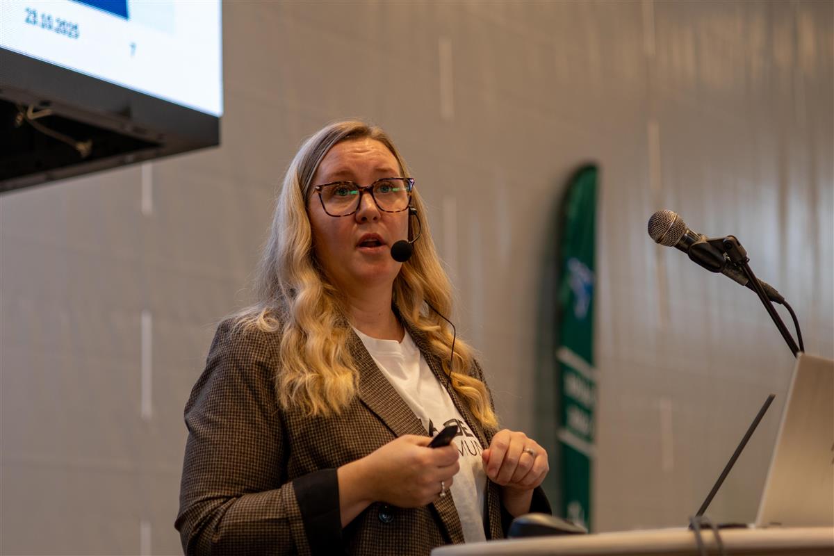 En kvinne står ved et podium foran en stor skjerm og snakker til et publikum. - Klikk for stort bilde