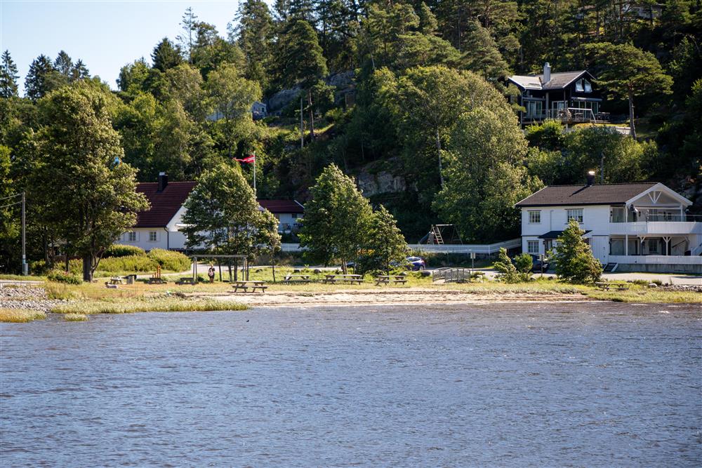 En strand sett fra lang avstand, masse trær bak stranden og noen hus. - Klikk for stort bilde