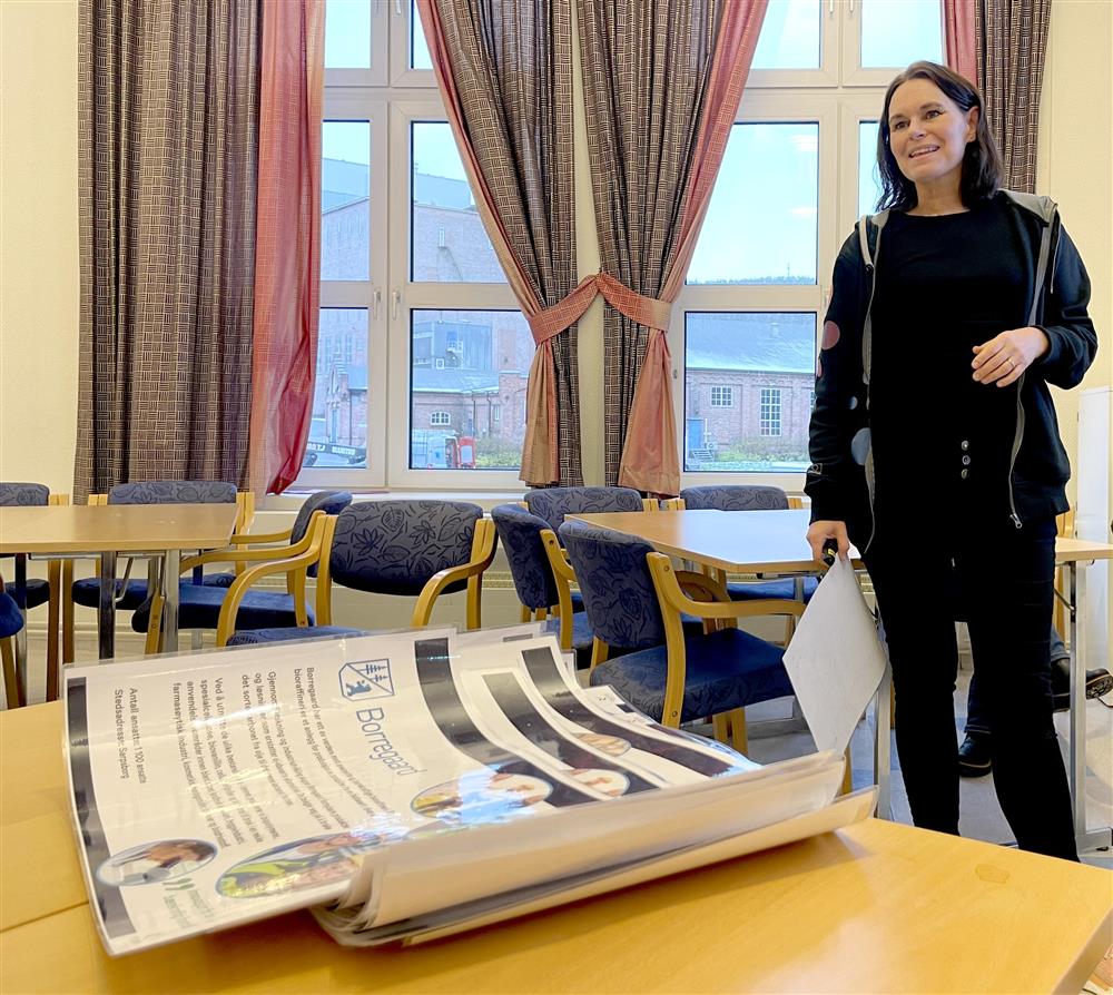 en dame med mørkt hår og mørke klær står ved siden av et bod hvor det ligger en haug med papirer - Klikk for stort bilde