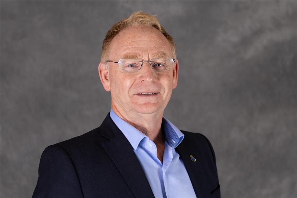 Mann med kort hår og lyse briller , har på seg en dressjakke med blå skjorte under, står foran en grå bakgrunn - Klikk for stort bilde