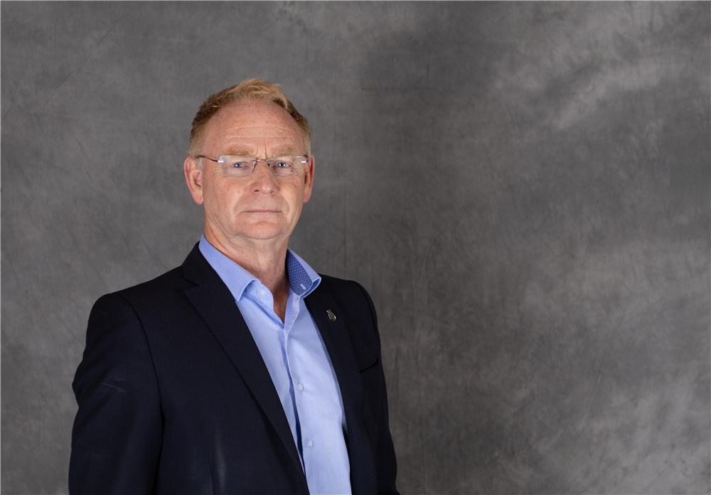 Mann med kort hår og lyse briller , har på seg en dressjakke med blå skjorte under, står foran en grå bakgrunn - Klikk for stort bilde