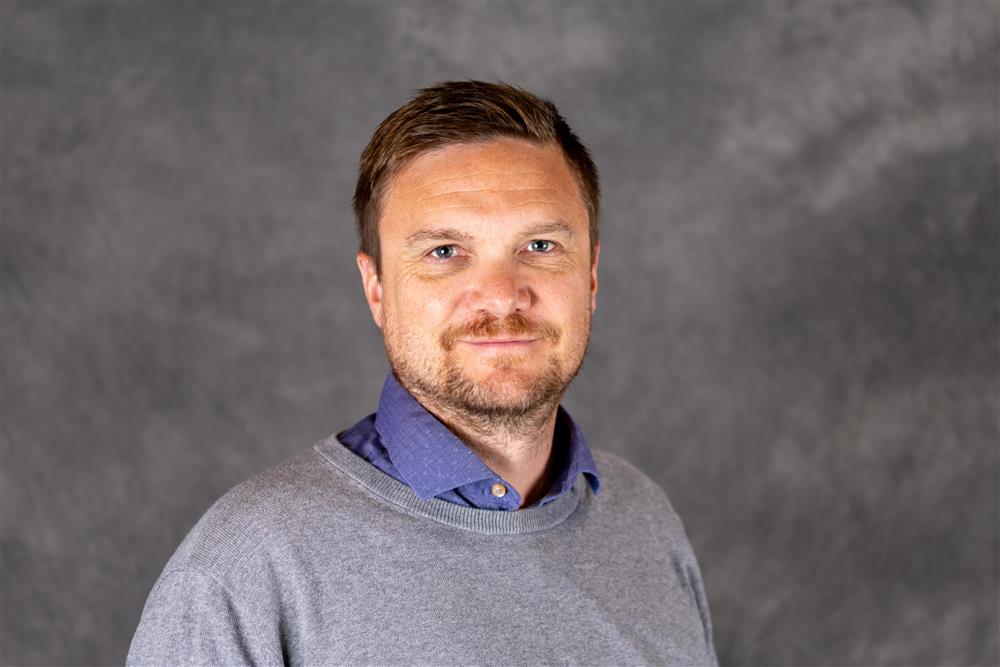 Mann med kort mørkt hår og grå blazer ser inn i kameraet - Klikk for stort bilde