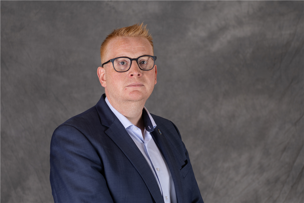 Mann med kort oransje hår og briller har på seg blå dressjakke foran en grå bakgrunn - Klikk for stort bilde