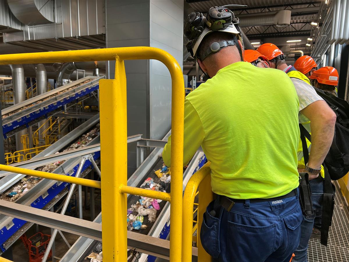 Personer med hjelm og arbeidsklær ser på sorteringsanlegg for avfall - Klikk for stort bilde